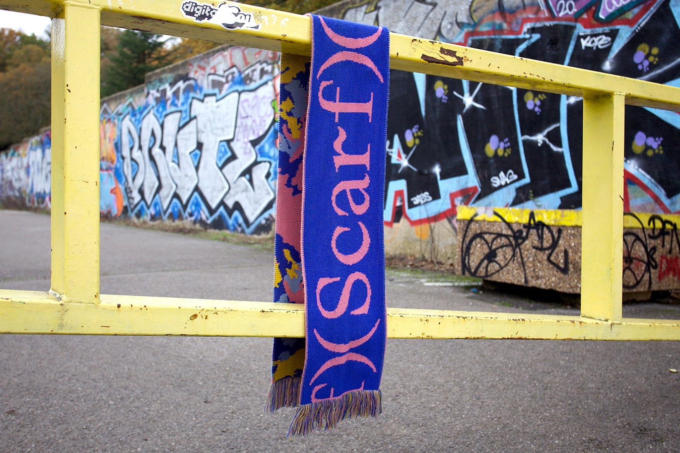 The final image of scarf has the scarf hanging from a metal gate, behind it is graffiti and urban settings
