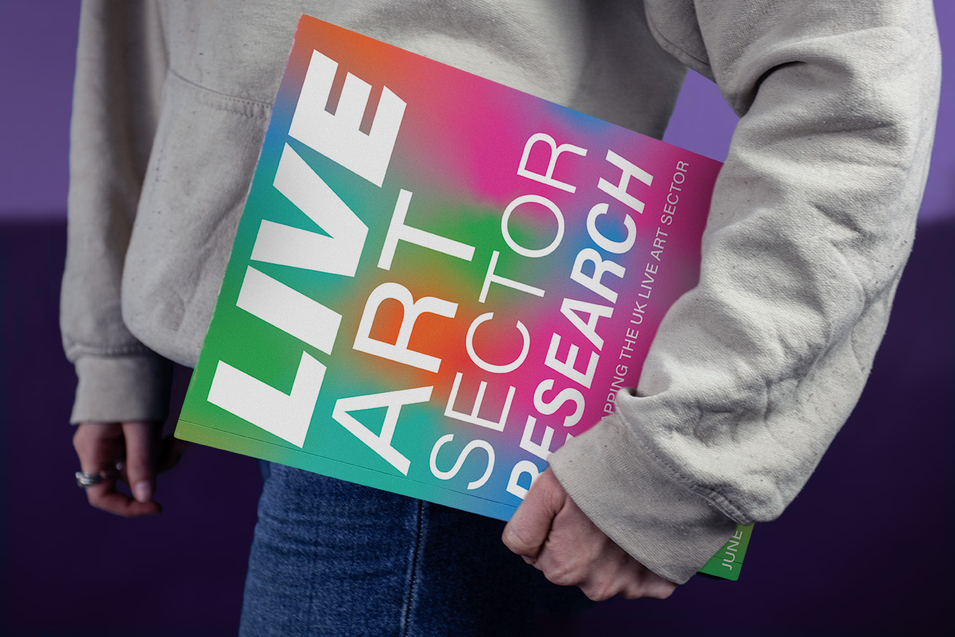alt: A woman holding the Live Art Sector Research book. The front cover uses multiple font weights and bright gradients to catch the eye