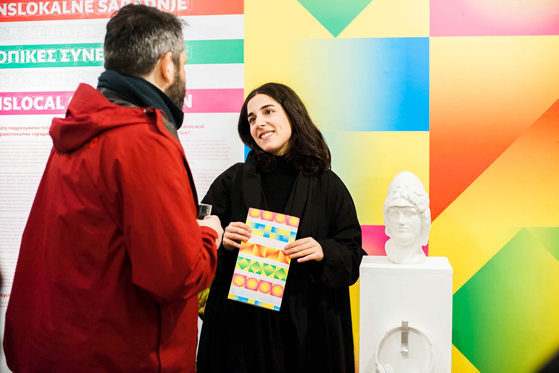 An image of one of the artists exhibiting within the Furtherfield Gallery space, in her hand is a Translocal Cooperation booklet. On the wall behind the artist has large geometric colourful shapes which are used throughout the Visual Identity.