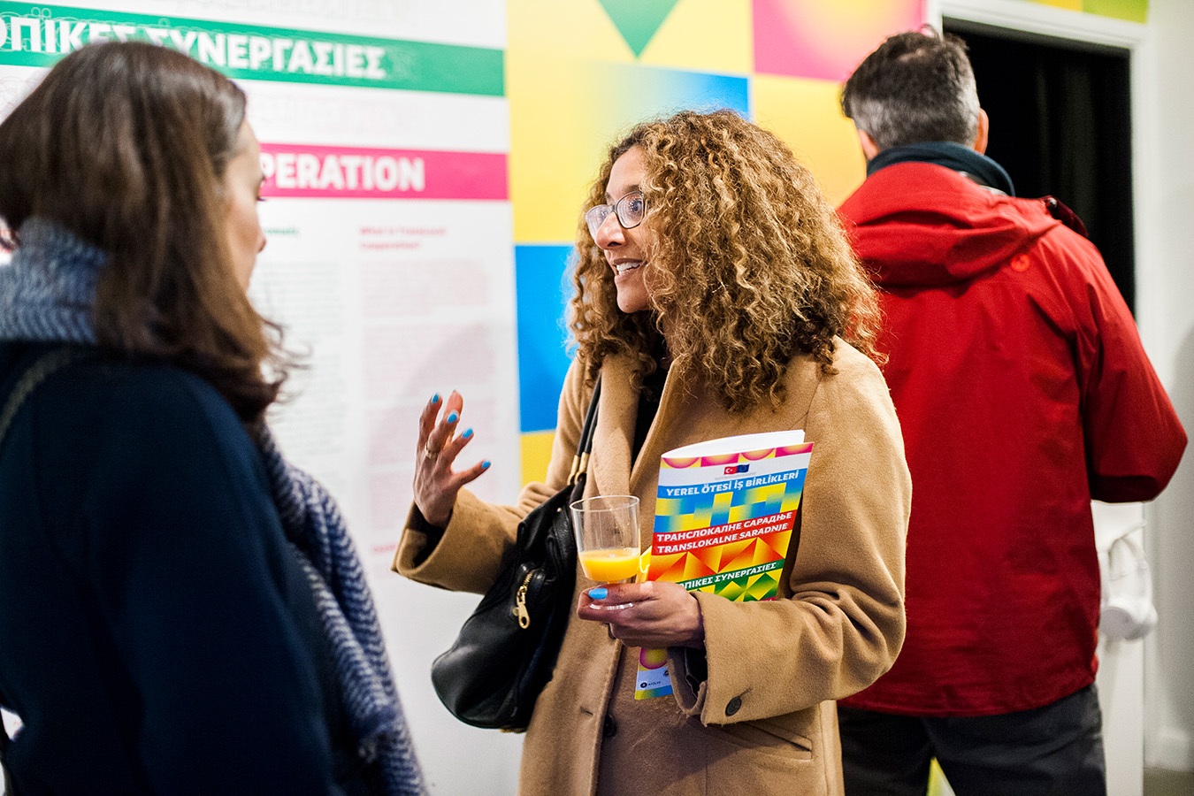 A member of the public holding the Trans Local Co-operation booklet, standing in front of the visual identity which has been applied in a large format wallpaper covering, the member of the public is talking and smiling to another during the exhibtion