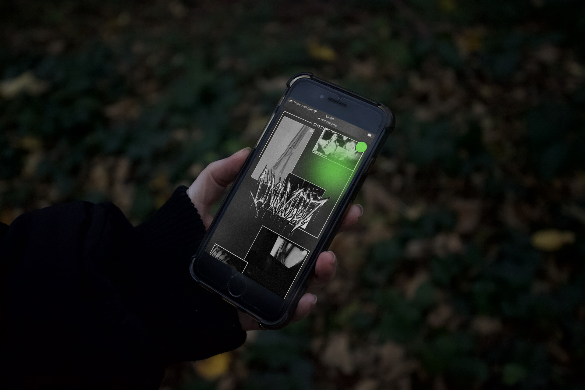 a Hand holding a phone in front of a forest floor, displayed on the phone is the Uninvited mobile site homepage