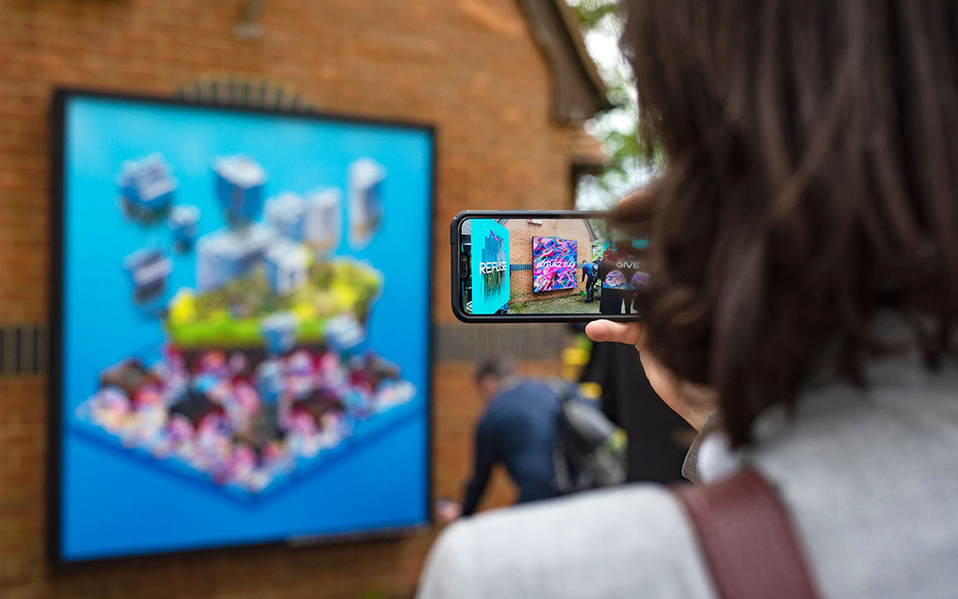 alt: A person holding a phone in front of a billboard on furtherfield gallery, through the phone you can see AR being using to view virtual artworks.
